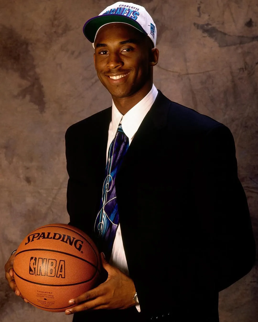 Young Kobe Bryant wearing a Charlotte Hornets cap and holding a basketball during the 1996 NBA Draft portrait.